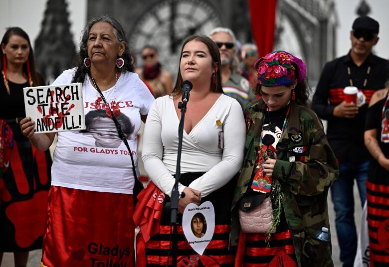 Three women stand at a microphone wearing red ribbon skirts.