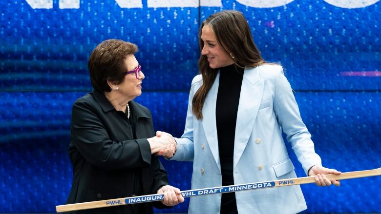 Two women shake hands while holding a hockey stick.