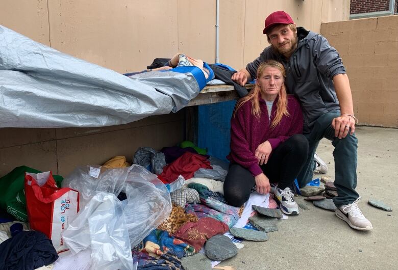 Tarps and blankets hang over a wooden shelf. A man and woman are crouched down near the makeshift tent. 