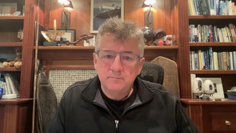 A man sitting in a chair with bookshelves in the background. He has grey hair and glasses.