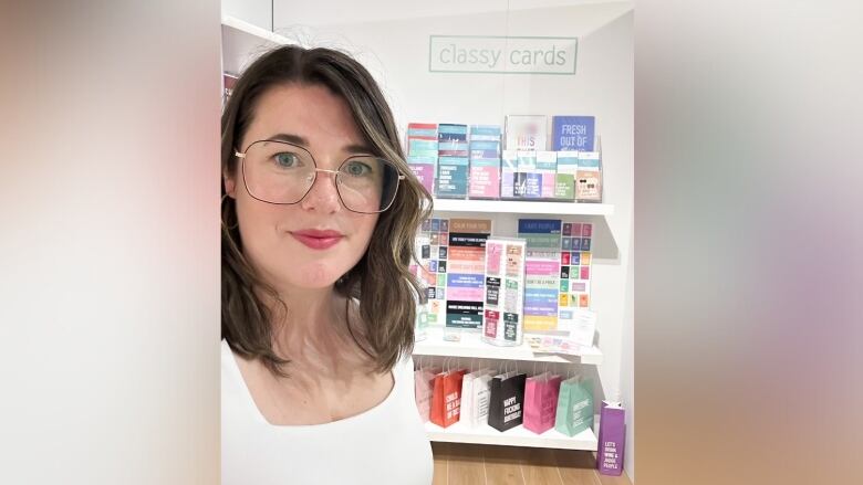 A woman wearing glasses takes a selfie in front of a wall display that holds cards, calendars and gift bags. 