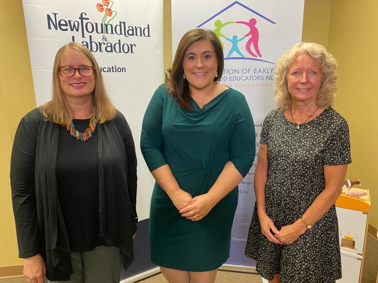 Three smiling women stand together in front of two banners for the Newfoundland and Labrador government.