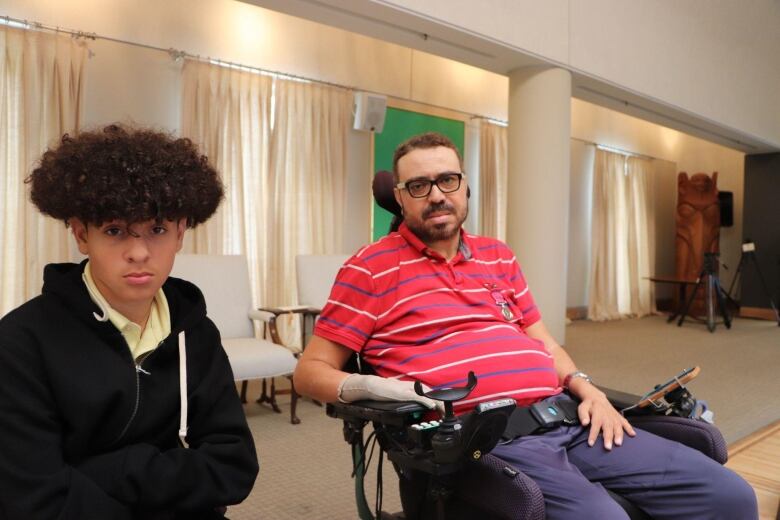 A man in a motorized wheelchair and red striped shirt sitting next to his son wearing a black hoodie