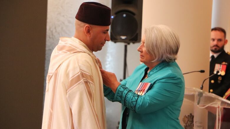 A man on the left stands as a woman on the right affixes something to his robe.