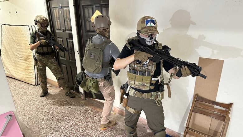 Three people in military gear move through an indoor space, guns drawn. 