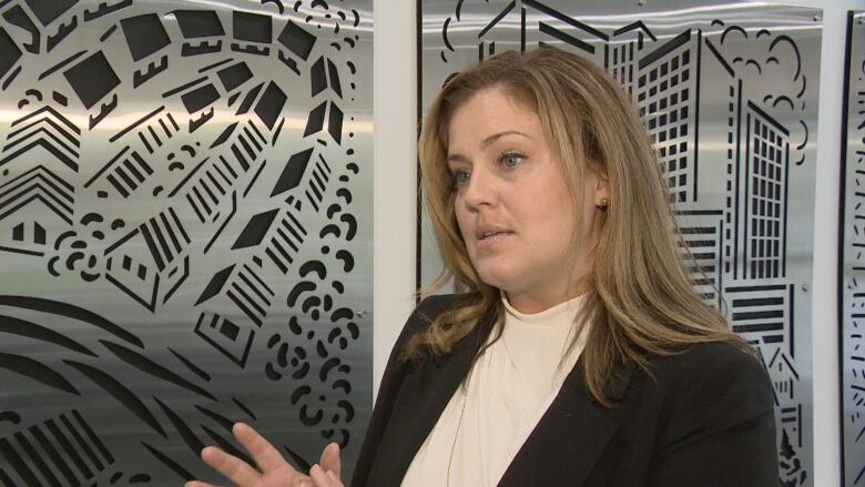 A woman with long blond hair and a black blazer stands in front of a silver art background