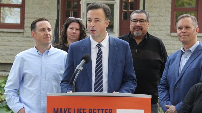 A man in a blue suit and tie speaks at a microphone, behind a podium, while flanked by a few other people.