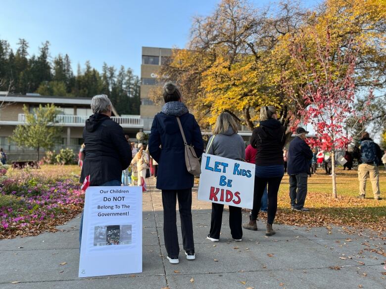 People stand with their backs to the camera with signs resting behind them. One sign reads 'Let Kids Be Kids'.