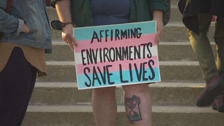 A protester holds up a sign painted in the trans pride flag colours with the text 'affirming environments save lives.' 