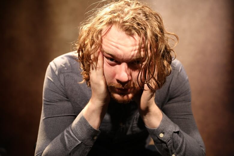 A man with shoulder-length straggly hair holds his face in his hands.