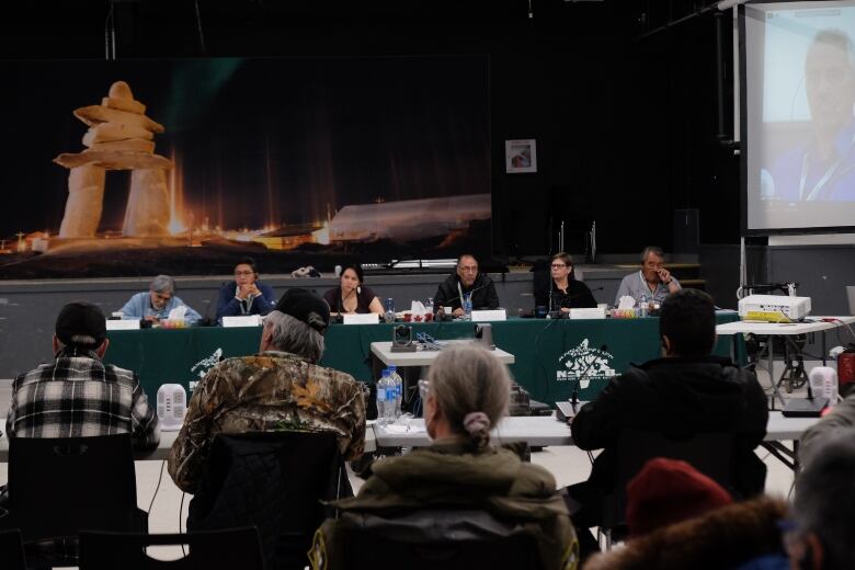 People sit at long tables with microphones inside a large gymnasium.