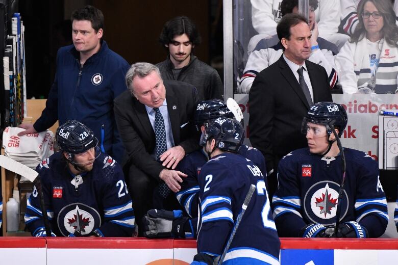 A man in a suit talks to a hockey player standing on the ice. Other players sit beside him.