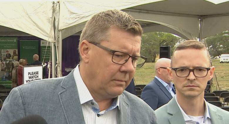 The Premier, wearing dark rectangle glasses, furrows his brow as he listens to a question. Another man, wearing dark rectangle glasses, stands slightly behind him. 