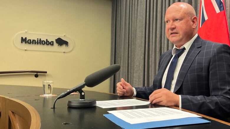 A man is seen speaking into a microphone while seated at a desk. On the wall next to him is a sign that says 