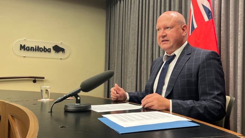 A man is seen speaking into a microphone while seated at a desk. On the wall next to him is a sign that says 