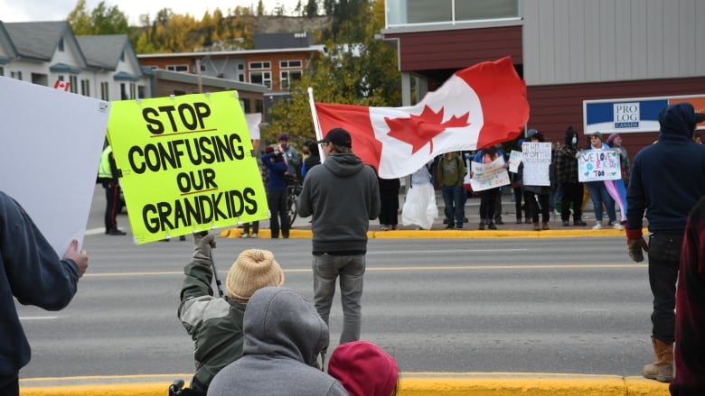 A crowd of people stands on the sidewalks on either side of city street, holding flags and signs.