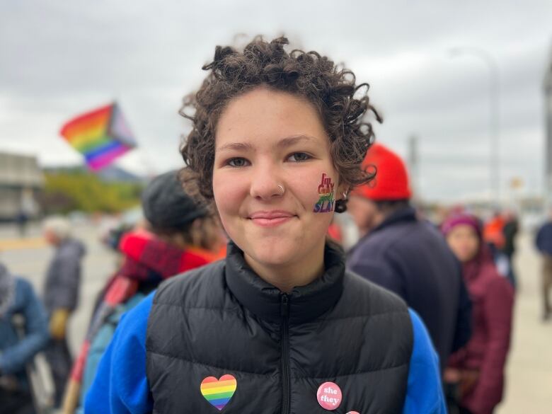 A portrait of a young woman on a city street with people visible behind her on the sidewalk holding rainbow flags.