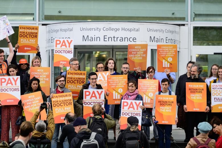 People holding orange placards and signs with slogans demanding better pay for doctors stand at the entrance of the University College Hospital in London, England. 