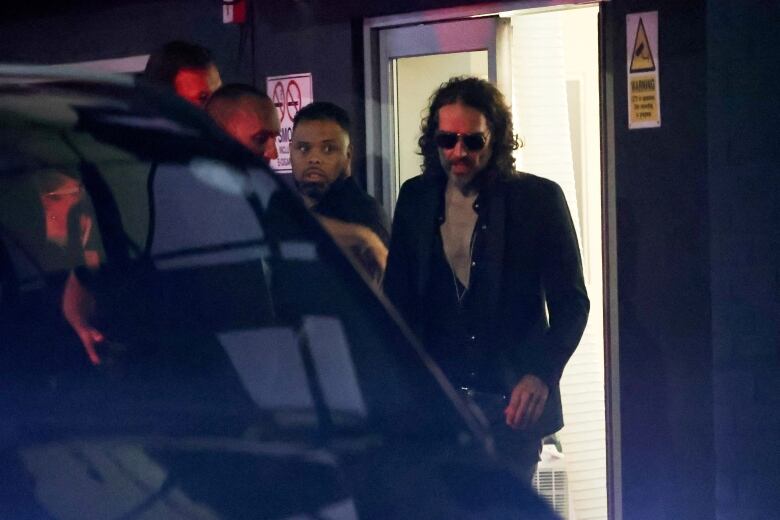 A man wearing sunglasses at night prepares to enter a vehicle.