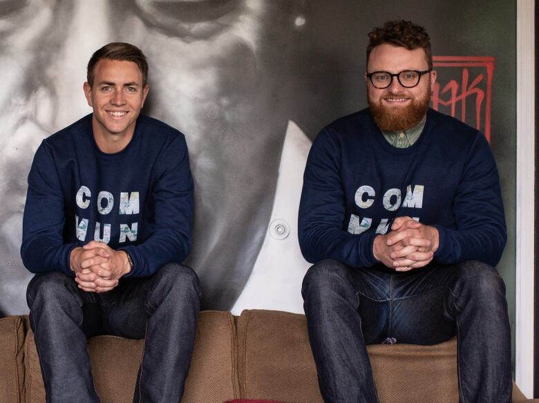 Two men sit with their hands clasped. They are wearing matching sweaters. 