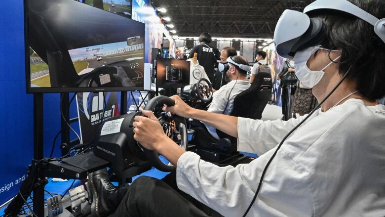 A man wears a virtual reality headset and controls a video-game version of a steering wheel to play a racing video game.