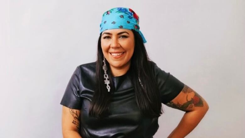 An Indigenous woman wearing a black short sleeve top, long silver earrings and a blue bandana over her hair, stands with one had on her hip, smiling at the camera. 