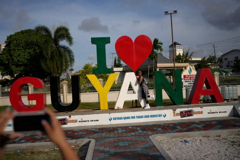 A woman poses by a sign saying 'I heart Guyana.'