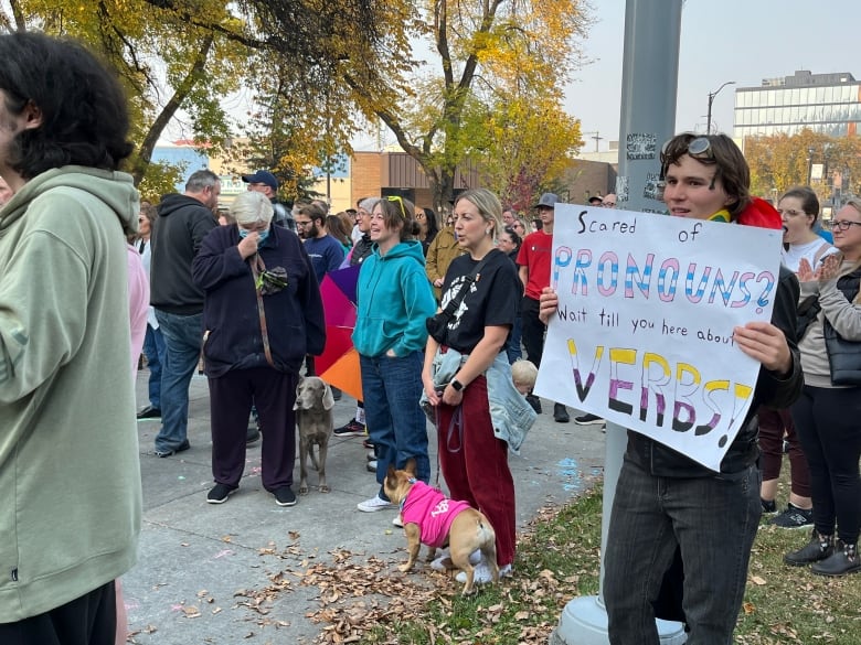 A person holds a sign that says scared of pronouns? wait til you hear about verbs