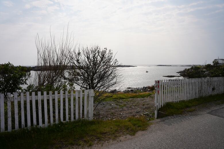 A white picket fence sits in front of a vacant lot, where a home used to stand. The water is very close to the land, and a boat sits on the water in the background.