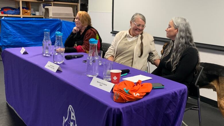 Three women sit behind a table with a purple tablecloth draped over it. A dog stands behind them.