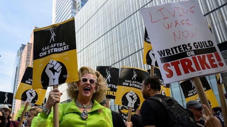 A pickecter in a green dress and wearing sunglasses holds a sign on a street, surrounded by other picketers.