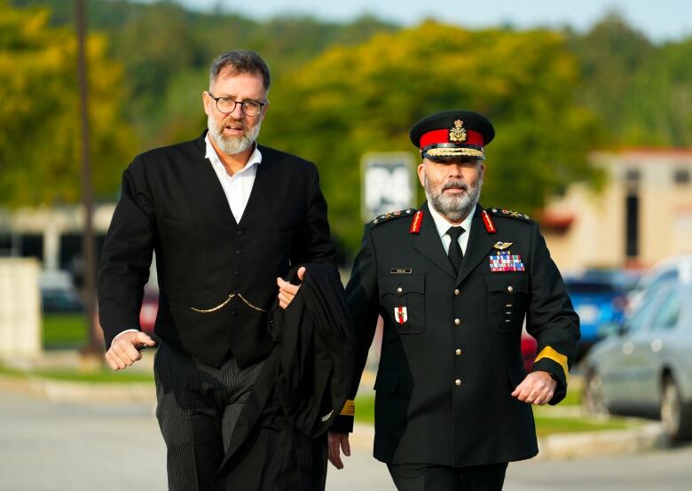 One person in legal attire and one in a military uniform walk along a road.