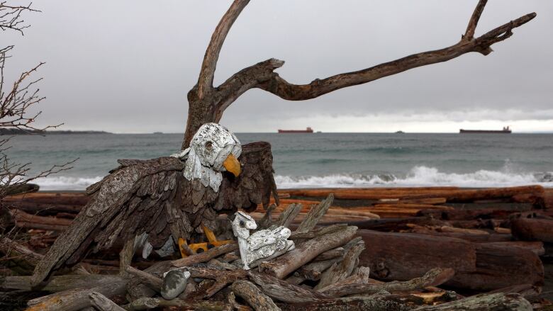 A driftwood sculpture of an eagle and chick with waves crashing onto a shore behind, under cloudy skies.