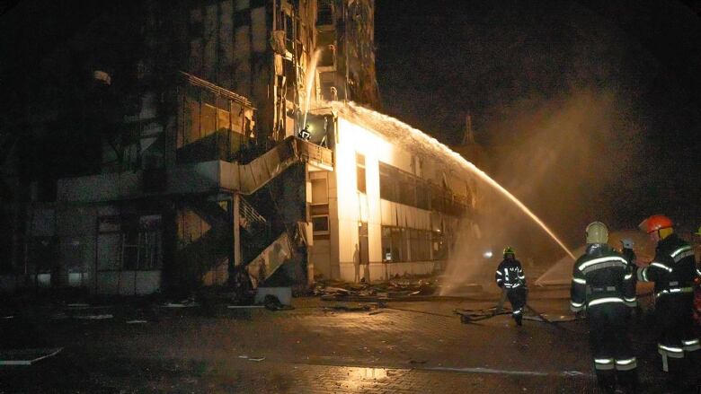 Peoplein black firefighter suits stand in the right corner of the frame aiming a hose at a damaged building at night. 