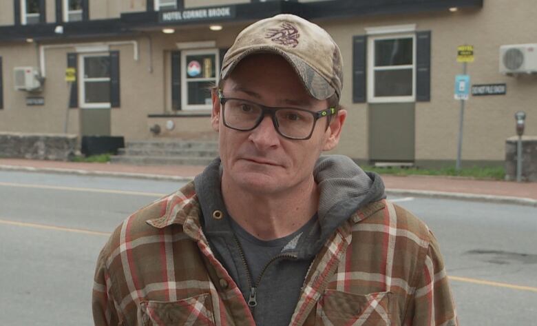 A man wearing a baseball hat, glasses and a plaid jacket stands in front of a beige and grey building. 