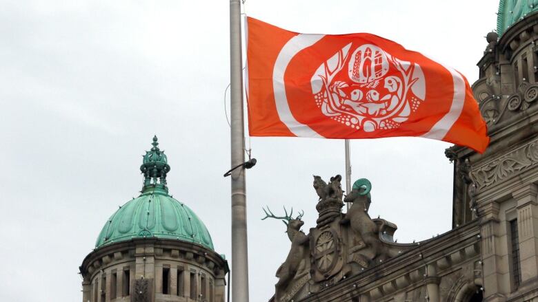 An orange flag with white Indigenous designs flies against a slate grey building with turquoise turrets.