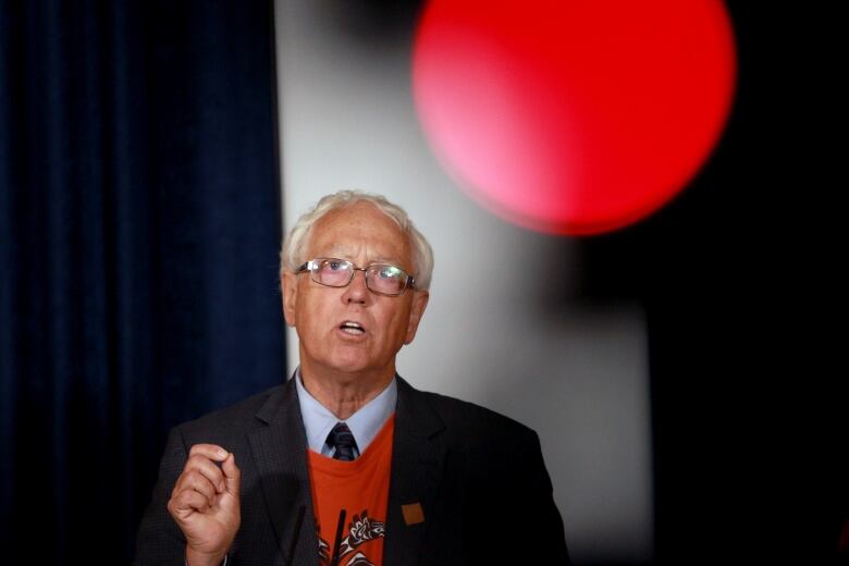 A white-haired caucasian man with glasses stands at a podium speaking. He wears an orange t-shirt with an Indigenous design on it over a dress shirt and tie. His mouth is open to speak.