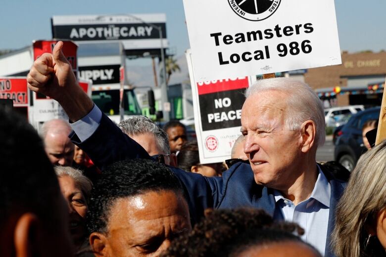 A man is shown in the middle of a crowd in a demonstration that includes signs, one which reads 'Teamsters Local 986.'