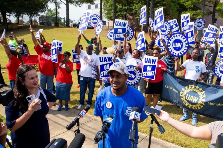 A man is shown standing in front of several microphones, with several more people behind him on a grassy area holding up signs that say, 'UAW on strike.'