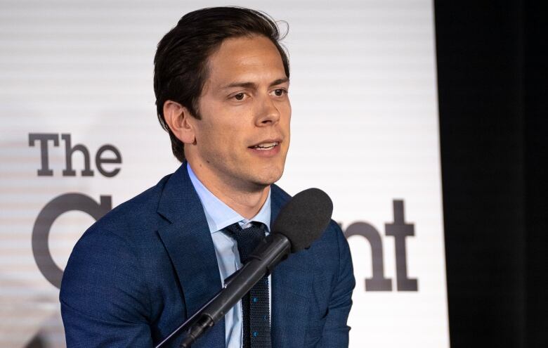 A man with short brown hair and a blue suit speaks into a microphone onstage at a forum. Behind him there is a sign that says, 'The Current.'