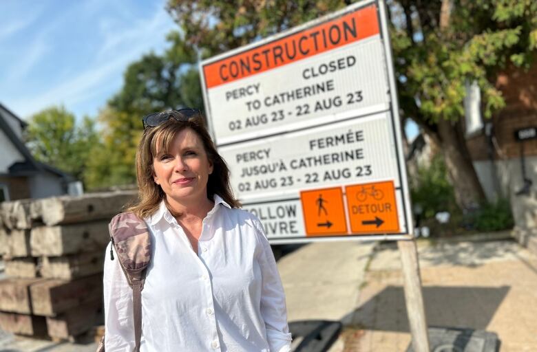  A woman with brown hair and white shirt stands in front of construction sign.