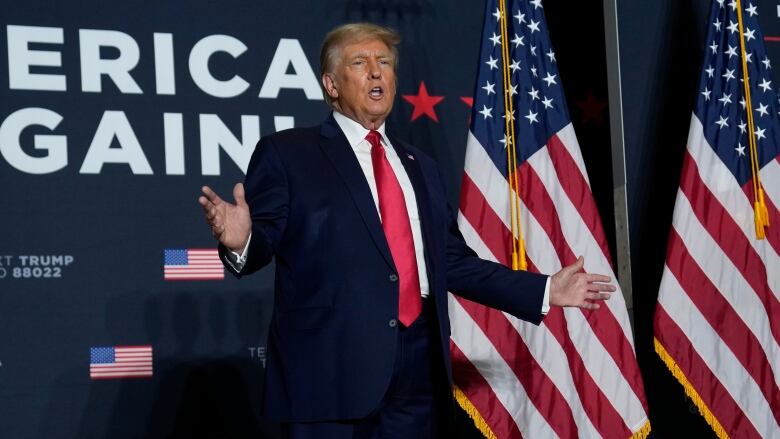 A man in a navy blue suit, white shirt and red tie stands with his arms extended in front of U.S. flags and a blue wall with the words 