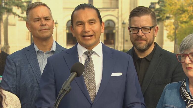 A man in a blue suit speaks at a microphone, surrounded by other people.