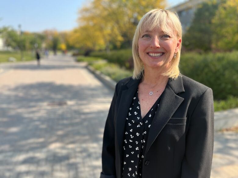 Woman with short blonde hair stands outside wearing a black suit jacket and a black shirt with a small white pattern. 