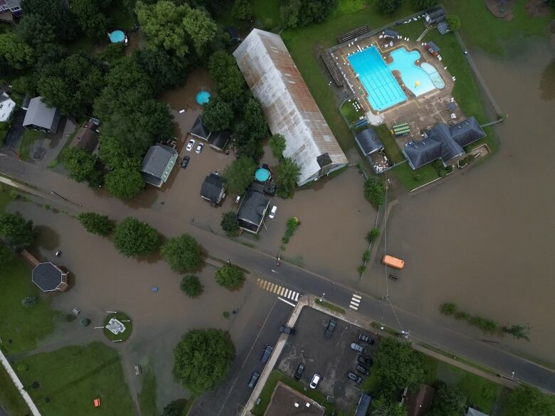 A drone shot shows brown water overflowing into the streets and lawns of a residential area. there's a bright blue swimming pool. 