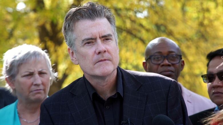 A man in a black suit speaks at a microphone to reporters off camera. 