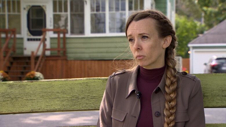 A woman looks off to the side. She wears her hair in a long braid. There's a green house behind her. 