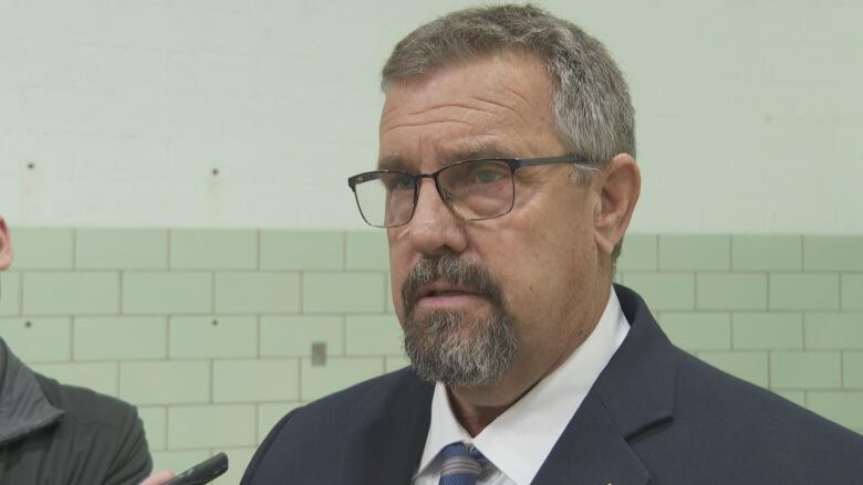 An unsmiling man with a grey beard and black-framed glasses