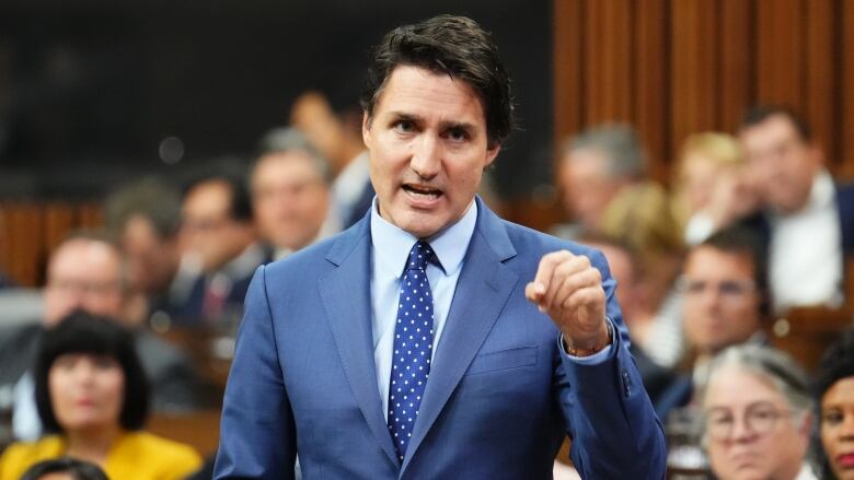 A man in a blue suit stands and gestures with his hand.