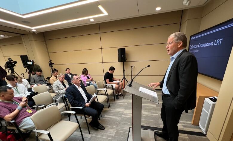 A man speaks in the front of a room full of people. A presentation is loaded onto a screen behind him.